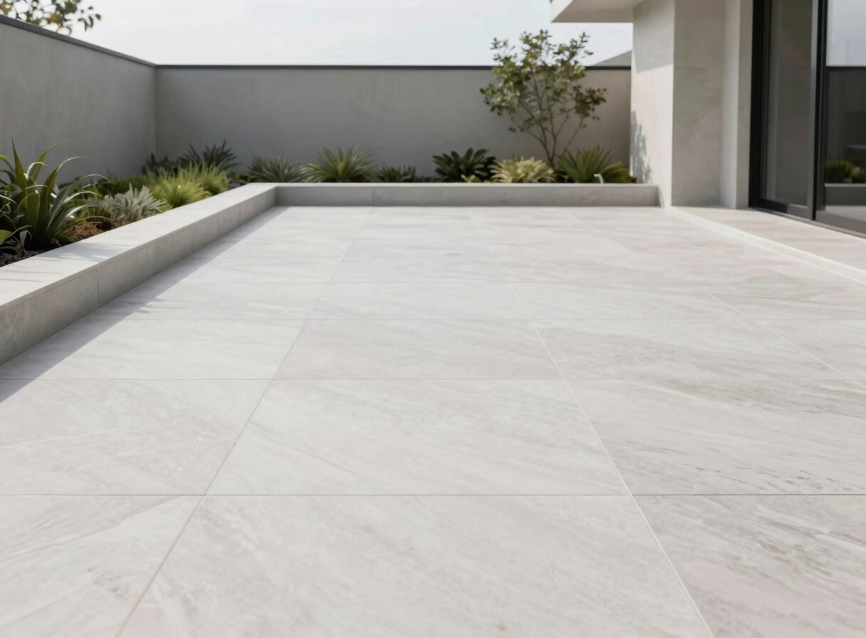Minimalist modern patio with large format porcelain slabs, clean borders, architectural landscaping in the background, soft overcast daylight